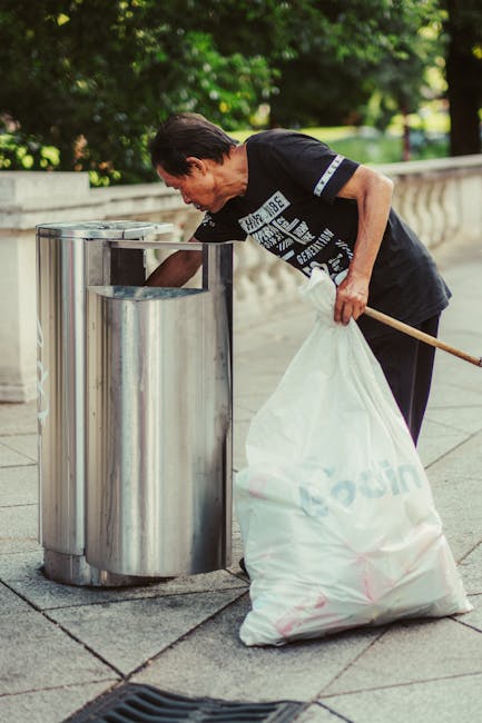 A middle-aged man with dark hair and a black T-shirt is seen using a metal litter bin located on a paved sidewalk in an outdoor urban setting. The man is slightly bent forward, depositing waste into the top opening of the cylindrical stainless steel bin, which has a polished finish reflecting the surroundings. He is holding a long-handled tool or stick in his right hand, and has a large white plastic bag filled with rubbish secured under his left arm. The background features lush green trees and shrubbery, with a stone balustrade running parallel to the sidewalk, suggesting a park or street in a city setting. The scene is well-lit, with natural daylight highlighting the details of the man’s clothing, the texture of the plastic bag, and the shiny surface of the waste receptacle. This image exemplifies a responsible waste disposal activity in an outdoor environment, aligning with private rubbish collection services offered by companies like House Clearance Mayfair, emphasizing proper on-site waste management and disposal practices.