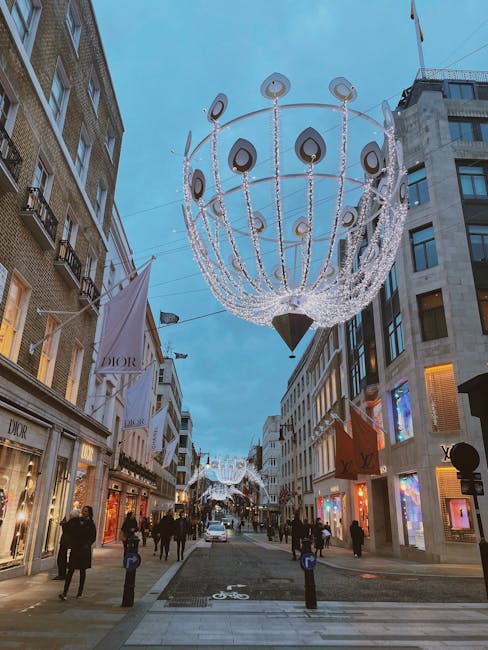 A busy shopping street during early evening with decorative street lighting in the form of a large, spherical chandelier-like installation hanging above. The chandelier features multiple circular elements with black and beige tones, connected by strings of bright white LED lights, creating a festive atmosphere. Flanking the street, luxury storefronts display illuminated logos and window displays, including the Dior boutique on the left, with its large glass windows showcasing fashion items. Pedestrians, some dressed in coats, walk along the pavement, indicating cool weather. The surrounding buildings are multi-story with modern and traditional architectural features, including stone and glass facades, and several windows lit from within. In the background, more decorative lights are visible, extending down the street, and a clear sky suggests early evening. This urban scene reflects an area that may be prepared for a festive or special event, with street lighting enhancing the atmosphere, which aligns with the concept of private or decorative street illumination rather than typical public or municipal lighting. House Clearance Mayfair specializes in handling waste from commercial and residential clearance, supporting the efficient disposal of waste generated in such busy urban environments.