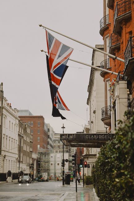 The image depicts a street scene outside the Claridge's hotel in Mayfair, with two Union Jack flags mounted on a pole extending from a building façade featuring balconies with ornate metal railings and greenery. The foreground shows a portion of the sidewalk with a hedge on the right side, and part of the hotel’s entrance sign reading 'CLARIDGE.' In the background, there are various multi-storey buildings, some with classic architectural details, and a few parked vehicles on the wet road, indicating recent rain. The overall atmosphere is overcast with diffused natural light, emphasizing a quiet urban environment. This scene relates naturally to waste management or rubbish removal themes, as the setting presents a typical upscale city street where private collection or on-site clearance services like those offered by House Clearance Mayfair may be employed to manage bulky waste or renovation debris in high-end properties.