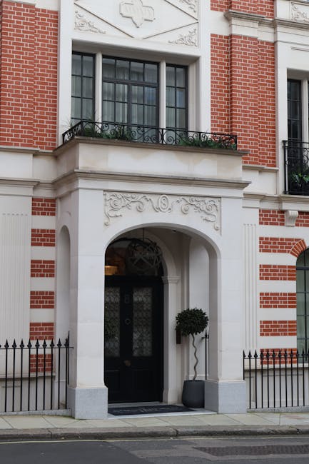 The image shows the front entrance of a historic-style building with a combination of red brick and white stone detailing. The doorway features a large, rounded arch with intricate carved patterns and a black door set within it, distinguished by decorative ironwork on the upper panel. On either side of the door, there are small, black metal fence posts connected by low rails along the sidewalk. Above the entrance, a window with a black frame and multiple panes is partially visible, topped with a decorative stonework panel that includes a cross symbol. A wrought iron balcony railing with floral motifs extends beneath the window, and a potted neatly trimmed shrub is positioned to the right of the door. The building’s facade combines traditional red brickwork with white stucco and carved detailing, suggesting a period architecture style often associated with upscale residential properties needing occasional cleaning or maintenance. House Clearance Mayfair, specializing in rubbish services, might be called upon for the safe removal of any accumulated debris or waste from such properties, emphasizing private waste handling or optional collection methods outside of local authority services. The scene is lit naturally, highlighting the detailed textures of brick, stone, and metalwork, conveying a well-maintained yet previously occupied residential entrance.