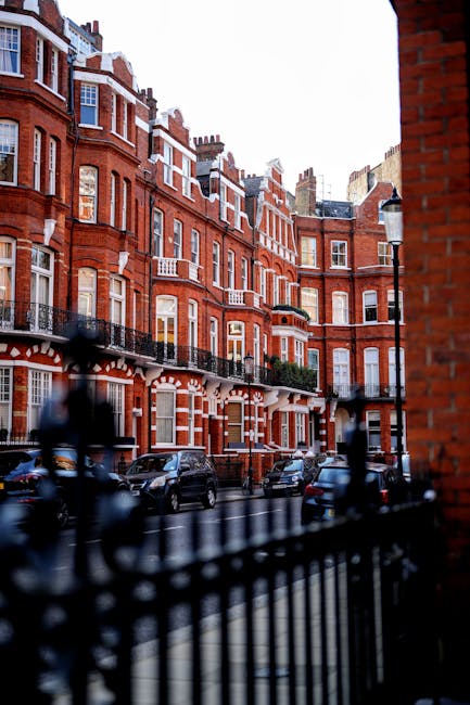 A street scene in a residential area featuring a row of traditional red-brick Victorian-style buildings with white window frames and decorative details. The buildings have multiple stories, some with small balconies and greenery visible on a few. Parked cars line the street alongside a black wrought-iron fence in the foreground, which is slightly out of focus. The street is illuminated by a classic-style street lamp and the sky suggests late afternoon or early evening lighting, with shadows cast on the facades. The environment appears quiet, with no visible pedestrians or movement. The overall scene reflects an urban setting where private property and street-level amenities are evident, aligning with the context of local rubbish clearance or alternative waste handling services like those offered by House Clearance Mayfair, helping facilitate on-site clearance of household waste from such residential properties.