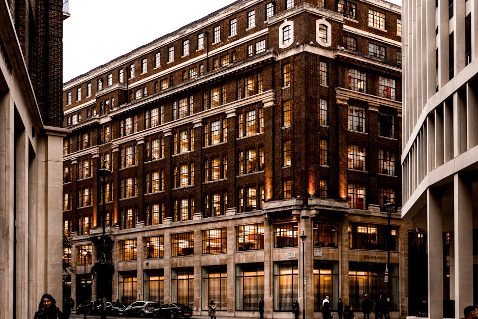 A multi-storey brick building with a classic architectural style, featuring numerous evenly spaced rectangular windows with black frames and glass panes reflecting the warm interior lighting. The facade is composed of dark reddish-brown bricks, with white horizontal decorative bands accentuating each floor. The lower part of the building has large ground-floor windows and a stone base, with a street sign attached to the corner indicating Mayfair location. Adjacent structures include a modern white building with vertical slats on the right, and a small corner lamp post nearby. The street scene shows parked cars at the curb and a few pedestrians walking along the pavement, captured during dusk or early evening, with the interior lighting of the building creating a warm glow contrasted against the overcast sky. The context suggests a city centre environment, where private or independent property management and potential waste removal services could be relevant for building maintenance or clearance activities, consistent with the themes of rubbish removal and estate clearance specialists like House Clearance Mayfair.