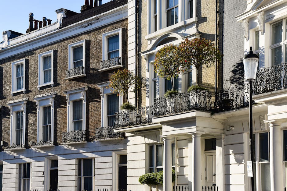 The image depicts a row of multi-story townhouses with classic London architectural features, including large sash windows with white frames and decorative lintels, and ornate wrought iron balcony railings. The buildings are constructed with brickwork in shades of dark brown and beige, complemented by white stucco detailing on the lower levels and around the entrance doors. In the foreground, a black cast iron street lamp stands tall beside the pavement, casting a shadow on the white facade of one building. Two small, well-maintained trees in pots are positioned on a balcony, adding greenery to the urban scene. The scene is illuminated by bright daylight, highlighting the contrasting textures of brick, metal, and foliage. The overall setting suggests a typical residential area that may require occasional private waste collection or rubbish removal services, as provided by companies like House Clearance Mayfair, particularly for maintaining tidy and clean exteriors in such prestigious areas.