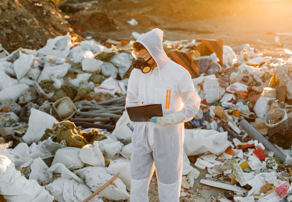 A person dressed in full white protective coveralls, including a hood, gloves, and a face mask with goggles, is standing amidst a large pile of mixed waste materials at an outdoor rubbish dumping site. The waste consists of white plastic bags, broken construction debris, Styrofoam, and assorted discarded items scattered across the uneven, dirt ground. The individual is holding a black clipboard and appears to be inspecting or assessing the rubbish, with sunlight casting a warm glow over the scene. In the background, there is a mix of earth and scattered debris, emphasizing the environment of waste disposal that may require professional private rubbish removal services such as those offered by House Clearance Mayfair, who handle various waste management tasks including hazardous and non-hazardous waste. The setting underscores the importance of proper waste handling and disposal, especially for bulky or potentially hazardous materials that may need special attention outside of standard municipal collection processes.