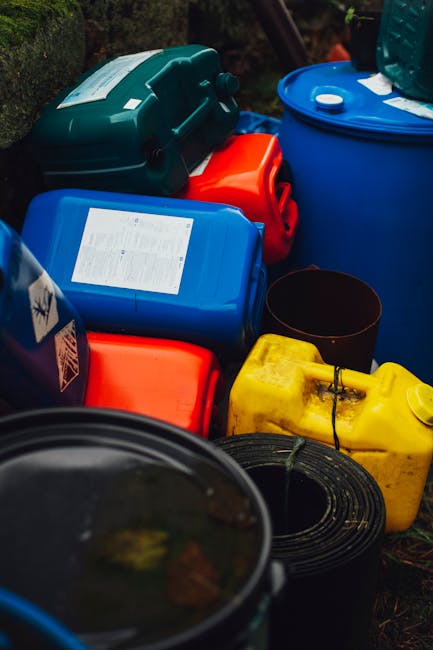 A close-up view of a pile of scattered rubbish behind a black metal fence, with crumpled paper, plastic bags, and food packaging in various colors including white, yellow, and red. The trash is resting on a patch of soil and fallen leaves, indicating an outdoor environment possibly near a residential area. The image showcases mixed waste materials, some of which appear light and flexible, contrasting with the rigid, dark metal bars of the fence in the foreground. The scene suggests an instance of improperly disposed waste, highlighting the importance of correct rubbish removal and private waste handling services like those offered by House Clearance Mayfair, especially in contexts where independent collection or on-site clearance may be necessary to maintain cleanliness and compliance with waste disposal obligations.
