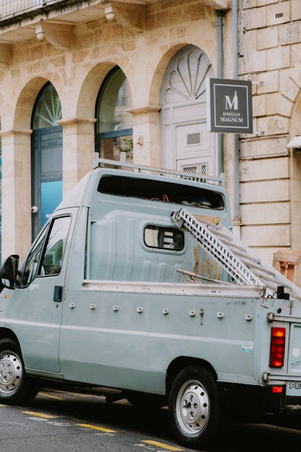 A light grey utility van parked on a city street in front of a historic stone building with arched windows and a decorative entrance. The van has a ladder secured on its side and appears to be used for waste or rubbish removal, with visible weathering and rivets along the bodywork. Behind the van, there is a black sign displaying a white letter 'M' and the words 'DORCHESTER MAGNUM' mounted on the building facade. The street surface is asphalt with a yellow dashed line, and the setting suggests an urban environment where private waste collection services like House Clearance Mayfair might operate as an alternative to local council rubbish pickups, especially for on-site clearance or specialised disposal.
