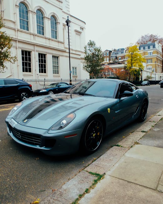 A metallic grey Ferrari sports car with a sleek, aerodynamic design is parked on a city street adjacent to a concrete sidewalk. The car features black alloy wheels with yellow brake calipers, a low profile, and smooth, curved body panels that reflect the surrounding environment. In the background, there are multi-storey residential buildings with yellow and white facades, large windows, and roof terraces, alongside a classic black lamp post and a few trees with early autumn foliage displaying yellow and green leaves. Several other parked vehicles, including a black SUV and a small silver car, are visible further along the street. The pavement appears clean, with a few fallen leaves scattered near the car and sidewalk, and the sky is overcast, providing soft, diffuse lighting. Though the focus is on the high-value vehicle, the scene subtly illustrates an urban setting where private or independent parking choices, such as on-site or alternative vehicle storage, might be considered, resonating with themes of personal disposal and disposal alternatives typically associated with rubbish removal and property clearance services provided by companies like House Clearance Mayfair.