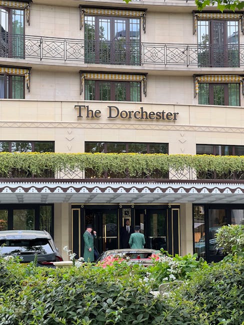 The exterior of The Dorchester hotel building, showing the entrance area with three staff members in green uniforms and a guest in dark clothing entering through black doors framed with gold accents. The facade features large windows with black and gold striped awnings and decorative ironwork balconies on the upper levels. A well-maintained green hedge runs along the front, partially obscuring the lower part of the building, and a silver car is parked near the entrance. The overall scene is set in daylight, with the building’s neutral-colored stone exterior contrasting with the greenery and the vehicle, reflecting a high-end hospitality setting possibly requiring private waste management or on-site clearance by companies like House Clearance Mayfair.