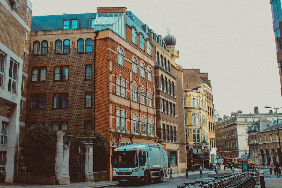 A white waste collection truck parked on a city street in front of a row of multi-storey buildings featuring a variety of architectural styles, including red brick and ornate facades. The truck is positioned near a gated entrance with stone pillars and greenery, possibly indicating a private courtyard or access point. Surrounding the truck, the street has barrier posts, bicycles, and street furniture, with additional vehicles and historical buildings visible in the background. The scene is captured during daylight hours with overcast or diffused lighting, highlighting the textures of the brickwork and the metallic surface of the waste collection vehicle. The setting suggests an urban environment where professional rubbish removal services, such as those provided by House Clearance Mayfair, operate to facilitate private waste disposal and on-site clearance, distinct from municipal rubbish collection. The image visually aligns with the context of independent waste handling in a cityscape, emphasizing the importance of professional rubbish services for maintaining cleanliness and order.