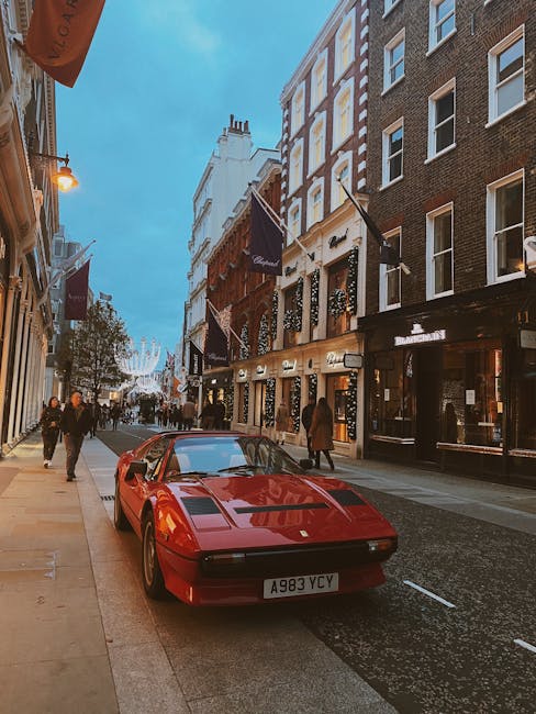 A red sports car with a sleek, low profile and smooth, glossy finish is parked on the left side of a busy city street, positioned partially on the pavement with its front facing toward the viewer. The car's angular design features rectangular pop-up headlights, a flat hood, and a European-style license plate reading 'A983 YCY'. The street is lined with multi-storey buildings featuring a mix of brick and painted facades, with large display windows showing interior lighting, and black and white awnings or signage above the entrances. On the upper floors, windows are evenly spaced and framed in white or dark trim. Several pedestrians are walking along the pavement, some dressed in casual clothing, and others in business attire, with a few individuals glancing toward the car. The atmosphere suggests early evening with a warm glow from streetlights and building exterior lights, and a clear blue sky overhead. The scene, captured during urban business hours, subtly evokes the context of private or alternative waste handling in a city environment, consistent with services like rubbish clearance or collection, often needed for premises such as shops or hotels on a commercial street. Occasionally, signs or flags attached to buildings hint at the presence of high-end retail or hospitality establishments, which may require specialised rubbish removal or on-site clearance solutions, as offered by local waste management services such as House Clearance Mayfair.