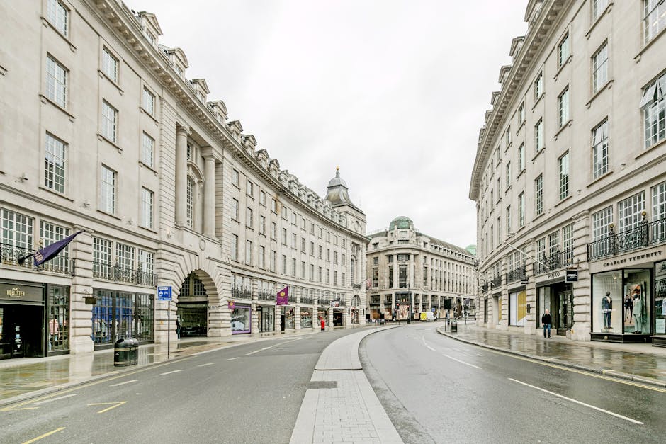 A city street scene featuring a mix of historic and modern buildings, with the main focus on a large, multi-storey brick building with ornate window frames and a rounded rooftop structure topped by a small dome. To the right, there are other buildings painted in light yellow and cream with architectural details, as well as modern glass and steel structures. In the foreground, a white waste collection vehicle with blue accents is parked on the street near a vintage stone entrance gate with decorative columns. The street is lined with metal bollards, and several bicycles are parked along the curb. The overall environment appears busy, with a variety of architectural styles and a typical urban atmosphere, highlighting the context of private waste management or rubbish collection services in a Mayfair location.