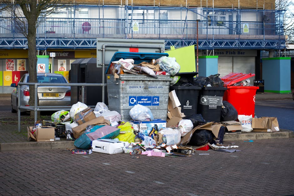 A large collection of rubbish and waste materials piled on a paved urban street corner, including numerous cardboard boxes, plastic bags filled with various debris, black and black-and-white trash bags, and loose paper and packaging materials scattered across the pavement. The waste is positioned next to a metal railing and partially obstructs a grey bin labeled for mixed paper and cardboard disposal, with the lid partially open and contents overflowing. In the background, a silver car is parked adjacent to the waste, with a building featuring shopfronts on the ground floor and scaffolding covering the upper levels. The scene is illuminated by natural daylight, suggesting daytime hours, and the surrounding environment indicates a commercial or residential area where independent rubbish collection, such as that offered by House Clearance Mayfair, might be necessary for efficient waste removal or site clearance.