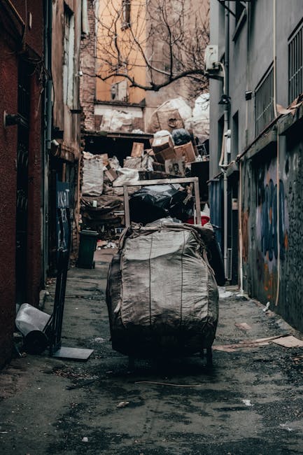 A large collection of rubbish and waste materials piled on a paved urban street corner, including numerous cardboard boxes, plastic bags filled with various debris, black and black-and-white trash bags, and loose paper and packaging materials scattered across the pavement. The waste is positioned next to a metal railing and partially obstructs a grey bin labeled for mixed paper and cardboard disposal, with the lid partially open and contents overflowing. In the background, a silver car is parked adjacent to the waste, with a building featuring shopfronts on the ground floor and scaffolding covering the upper levels. The scene is illuminated by natural daylight, suggesting daytime hours, and the surrounding environment indicates a commercial or residential area where independent rubbish collection, such as that offered by House Clearance Mayfair, might be necessary for efficient waste removal or site clearance.