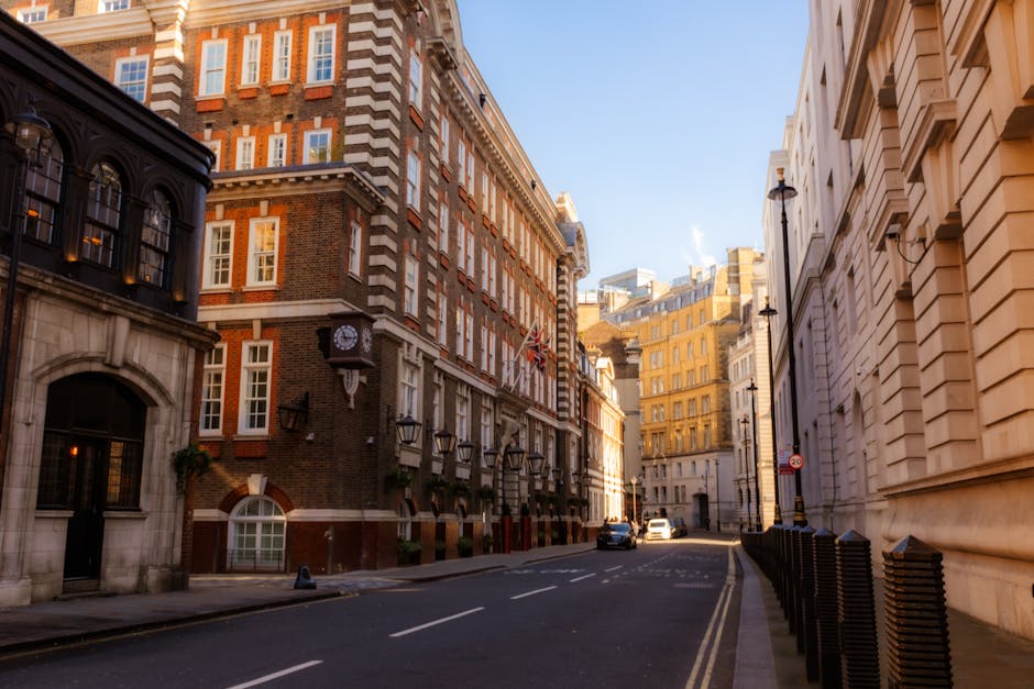 A quiet city street scene in Mayfair, showing a row of historic, multi-storey buildings with ornate architectural details and a mix of brick and stone facades. The building on the left features dark, polished window frames with glass panes and decorative lantern-style wall-mounted lights beneath a brick archway. The building has a small, white-framed, rounded window near the ground floor and a traditional clock mounted on its corner, with the clock face visible and showing the time. On the right, a cream-colored building with smooth stone exterior and rectangular windows extends into the distance, with black street lamps mounted along the sidewalk. The street is paved with asphalt and marked with a central double yellow line. Parked cars are visible further down the street, and the scene is illuminated by natural daylight, with a clear blue sky overhead. The area appears clean and well-maintained, consistent with urban street clearance and waste management practices. Occasionally, small plants and decorative elements are seen alongside the building facades, reflecting the tidy appearance typical of Mayfair's streetscape, while the scene captures the quiet atmosphere suitable for professional rubbish removal or private waste handling services by companies like House Clearance Mayfair.