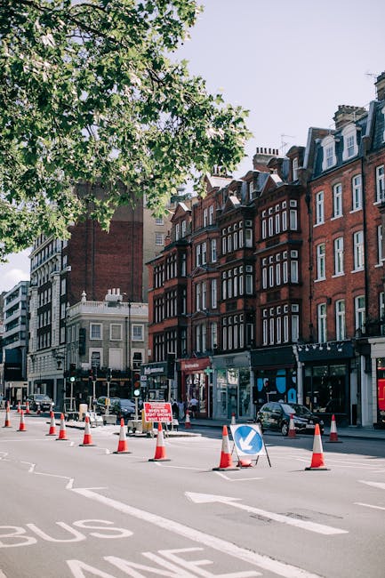 The image depicts a busy street scene in an urban area with a row of red-brick buildings featuring multiple windows and chimneys, typical of traditional London architecture. In the foreground, there is a section of the road with orange and white traffic cones blocking part of a lane, accompanied by a blue directional arrow sign indicating a diversion. To the left, a line of vehicles, including a bus and some cars, are positioned along the street, with several traffic lights visible at an intersection. A large green tree with leafy branches extends from the upper left corner, partially shading the scene and adding natural contrast to the cityscape. The background includes a mix of modern and older buildings, some with flat rooftops and chimney stacks. The scene is illuminated by natural daylight, with a light sky overhead, creating a clear and neutral atmosphere. This setting may relate to ongoing street or pavement work, with House Clearance Mayfair offering private waste removal or on-site clearance services to manage rubbish generated during urban refurbishments or street projects, reflecting the importance of alternatives to standard municipal waste disposal for local residents and businesses.